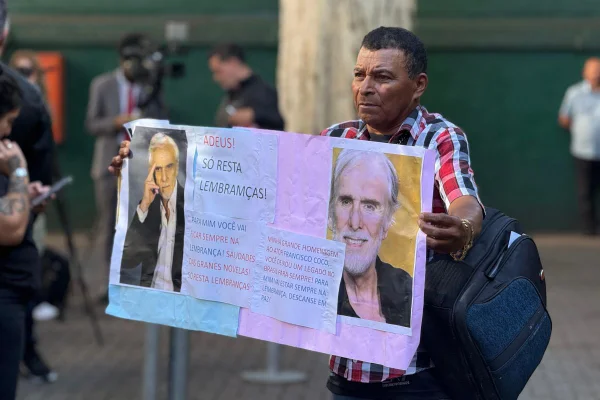 Imagem colorida mostra homem pardo vestindo camisa xadrez. Ele segura um cartaz com fotos do ator Francisco Cuoco - Metrópoles