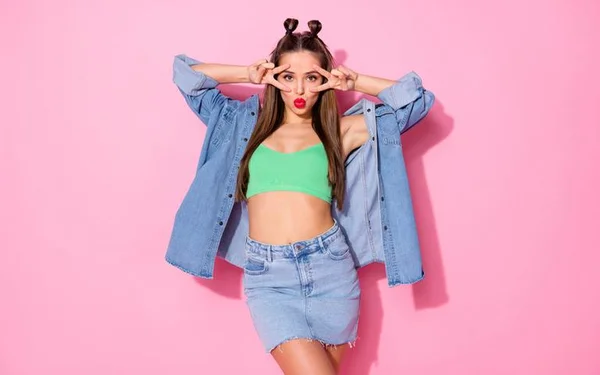 Mulher com cropped verde, saia e jaqueta jeans, fazendo uma pode com as mãos em um fundo rosa