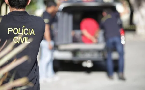 policial civil é fotografado de costas, ele usa o uniforme da corporação. ao fundo, um homem é colocado dentro de uma viatura da polícia