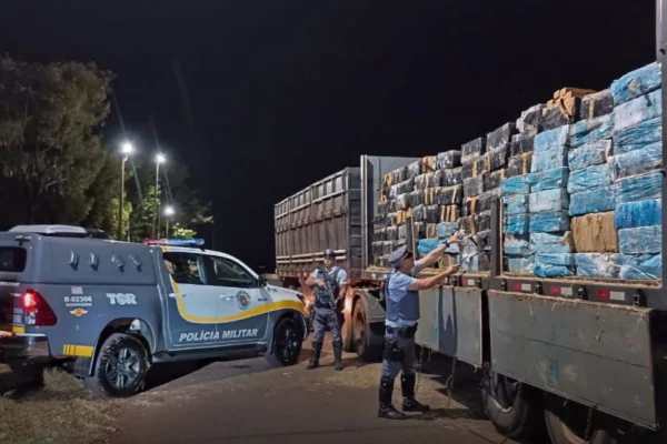 Imagens coloridas mostram policiais militares descarregando da caçamba de uma carreta uma carga de maconha apreendida em fardos de feno