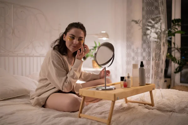 Jovem mulher sentada em cama fazendo skincare no rosto - Metrópoles