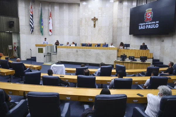 Imagem colorida mostra plenário com parede de mármore branco ao fundo e uma grande cruz no meio - Metrópoles