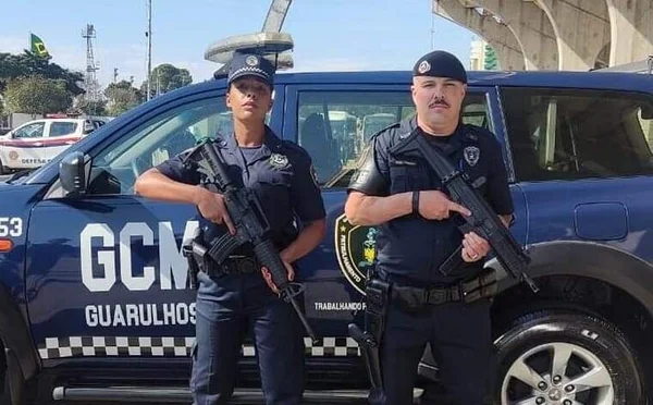 Em foto colorida Guarda Civil Metropolitana feminina segura fuzil, do lado esquerdo da imagem. Ao lado dele um GCM masculino, também empunhando um fuzil. Atrás deles, uma viatura azul escura da GCM de Guarulhos, Grande São Paulo - Metrópoles