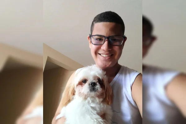 Foto de jovem com óculos, camisa branca e cabelo curto. Ele está com cachorrinho no colo
