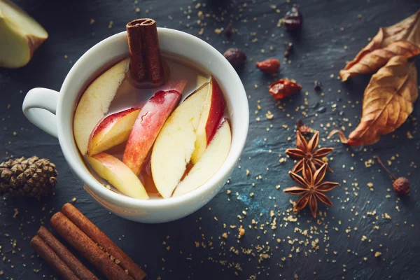 Foto mostra xícara de chá com fatias de maça, um pau de canela e açafrão ou cúrcuma espalhados em pó ao ser redor. Chá de maçã com canela