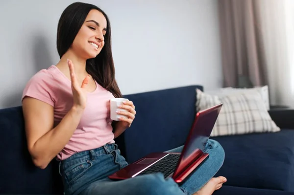 jovem-alegre-sentar-no-sofa-e-acenar-com-a-mao-ela-sorri-e-olha-na-tela-do-laptop-modelo-segura-lo-na-soneca-ela-tem-um-copo-branco-de-bebida_152404-4108