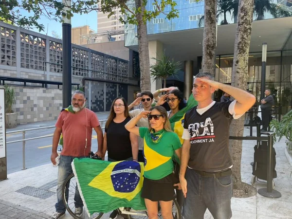 Imagem colorida mostra apoiadores do ex-presidente Jair Bolsonaro na frente do hospital onde ele está internado. O grupo segura uma bandeira do Brasil e um deles bate continência - Metrópoles