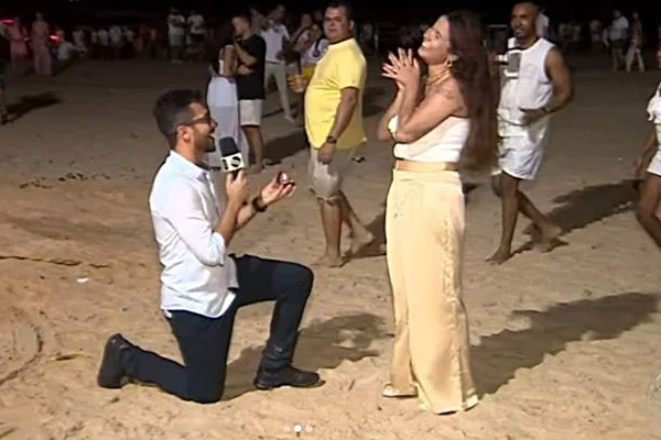 foto colorida de homem ajoelhado na areia segurando microfone e anel de noivado enquanto mulher sorri em pé - metrópoles