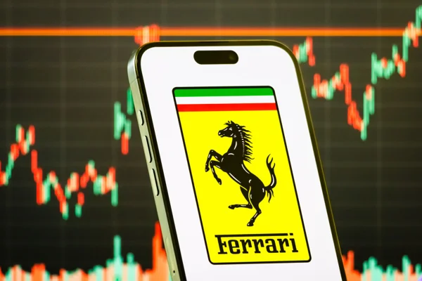 Imagem de celular exibindo a logomarca da Ferrari e, ao fundo, painel com o desempenho de ações negociadas na Bolsa de Valores - Metrópoles