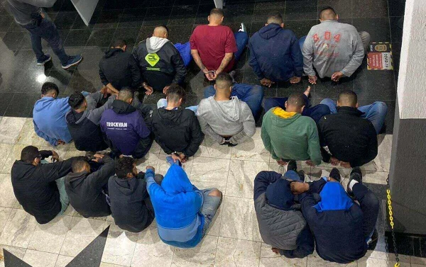 Em foto colorida 19 criminosos sentados no chão, de costas para a câmera - Metrópoles