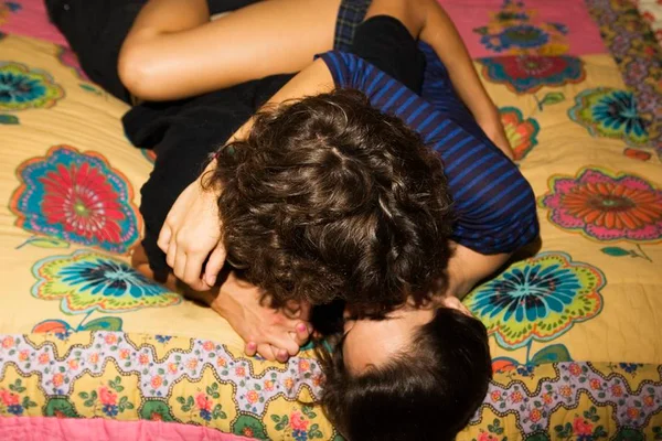 Foto colorida de dois jovens deitados em uma cama com lençol colorido abraçados e se beijando - Metrópoles