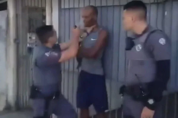 Imagem colorida mostra policial militar (PM) jogando spray de pimenta em um homem negro parado e imobilizado pelo pescoço - Metrópoles