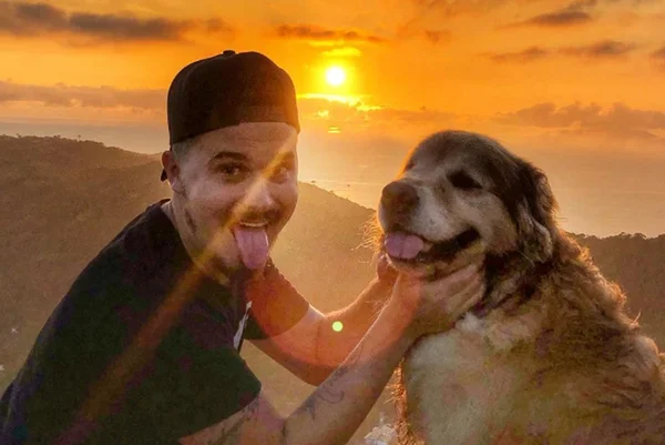 Foto colorida de um homem de boné ao lado de um cachorro na praia