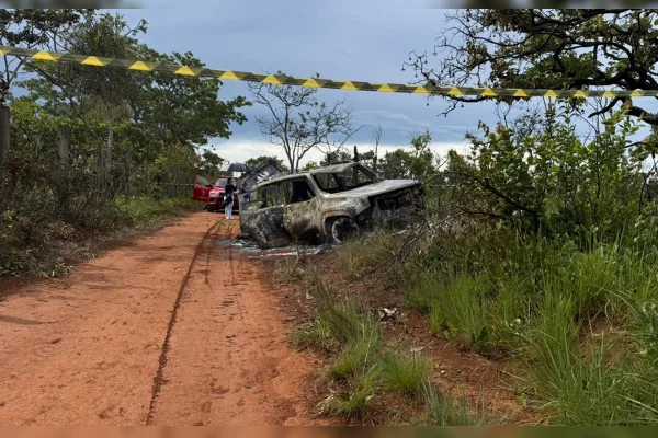 Imagem colorida de uma estrada de terra com mato dos dois lados; à frente, um carro branco, carbonizado; e uma fita em amarelo e preto isolando o local