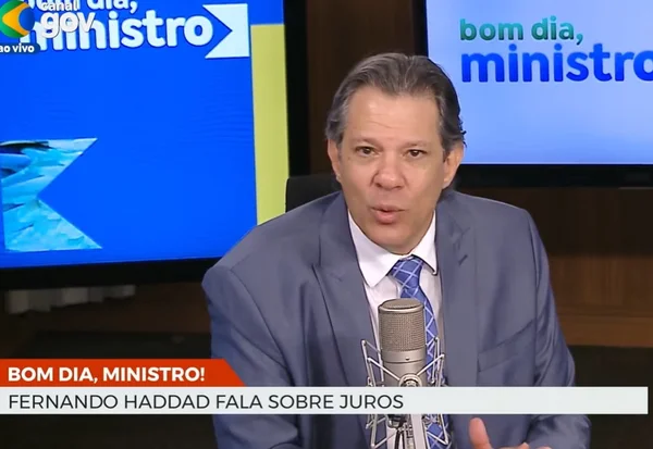 Imagem do ministro da Fazenda, Fernando Haddad, concedendo entrevista. Ele veste terno azul claro e fala em um microfone. Ao fundo, uma televisão com a imagem do logotipo do programa "Bom dia, ministro"