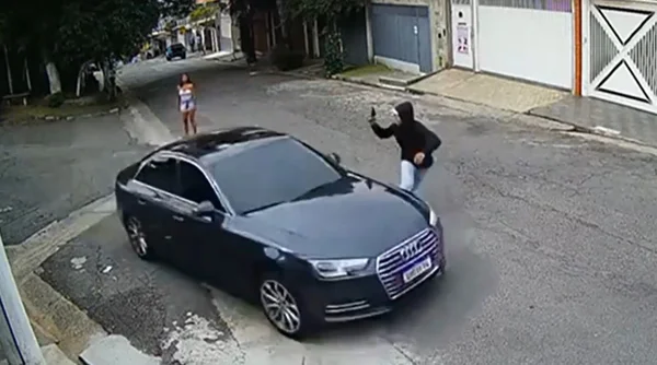 Imagem colorida mostra um homem de blusa preta com capuz apontando uma arma a um carro de luxo de preto; atrás deles, uma mulher vestida de branco e short jeans, aparentemente a isca para o chamado "golpe do amor" - Metrópoles