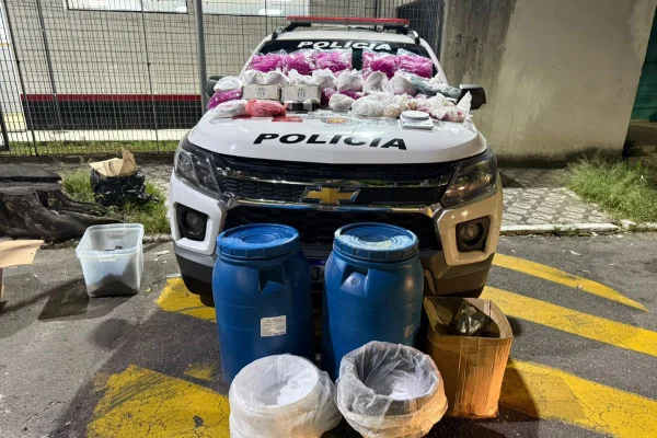 Imagens coloridas mostram viatura da Polícia Militar com pacotes de drogas no capô, tonéis azuis na frente do veículo