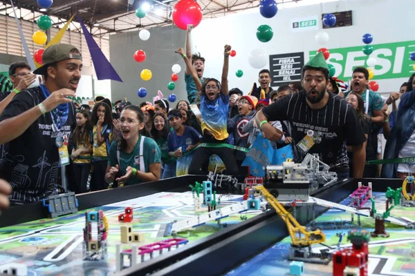 painel com maquetes e estudantes ao fundo comemorando