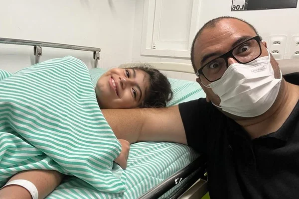 Foto de menina deitada em cama de hospital e homem de máscara do lado - Metrópoles