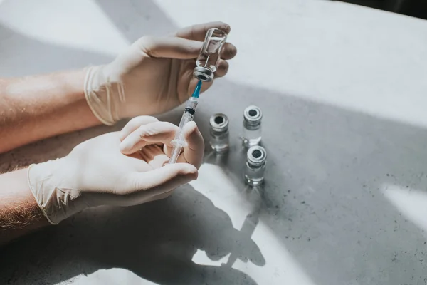 Uma mão com luvas de látex segura um frasco com líquido transparente e uma seringa. O profissional de saúde usa a seringa para extrair o medicamento do frasco de vidro. Metrópoles