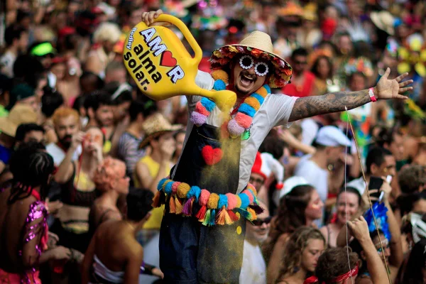 Homem está segurando um regador de planta e usando adereços coloridos durante o carnaval do Rio de Janeiro. Ele está usando óculos escuro, chapéu de palha e sorrindo pra foto - Metrópoles