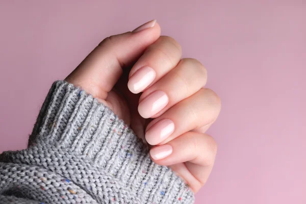 Mão feminina em um suéter de tricô cinza com unha nude rosada, sobre fundo rosa