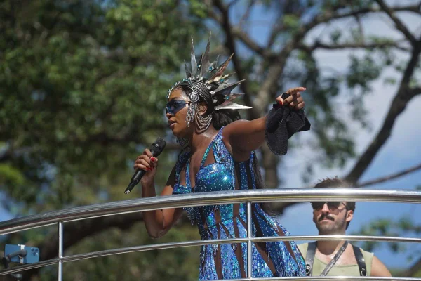 Imagens coloridas mostram cantora Iza em pé, microfone na mão, cantando durante apresentação sobre trio elétrico, ao fundo e céu azul entre nuvens e árvores