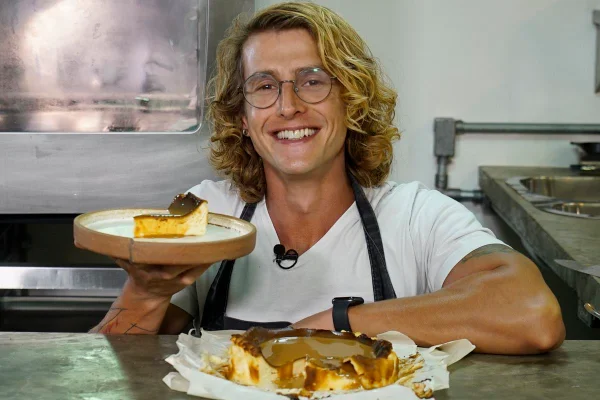 O cozinheiro André Rochadel sorri para a câmera com um prato na mão, mostrando uma fatia de cheescake basco. Na sua frente, o restante da torta - Metrópoles