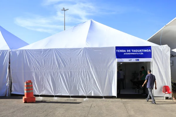 Foto colorida de tenda branca em estacionamento. Em uma placa azul sob a porta, está escrito: Tenda de Taguatinga