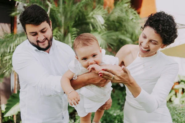 Imagem mostra um casal vestido de branco balançando um bebê - Metrópoles