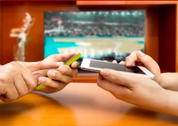 Fotografia colorida mostrando duas pessoas de lados opostos de uma mesa jogando nos celulares com uma TV ligada ao lado deles-Metrópoles