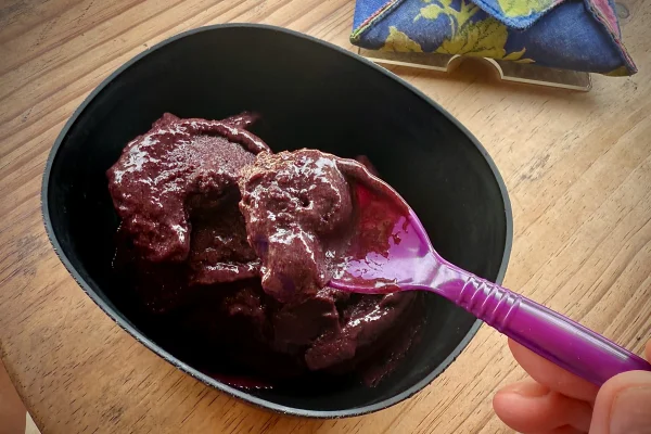 Foto colorida de tigela preta com açaí de cor roxa ao lado de mão masculina segurando uma colher - Metrópoles.