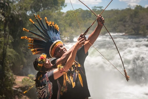 cacique de tribo indígena e esposa com arco e flecha em frentea a cachoeira - metrópoles