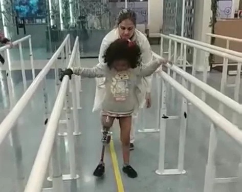 Menina com prótese na perna direita caminha com auxílio de uma mulher entre barras de apoio - Metrópoles