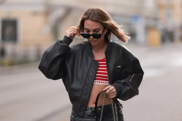 Mulher branca e jovem, de cabelo liso loiro, posa para foto na rua. Ela usa um top cropped listrado, branco e vermelho; calça jeans preta; jaqueta bomber preta e óculos escuros. - Metrópoles