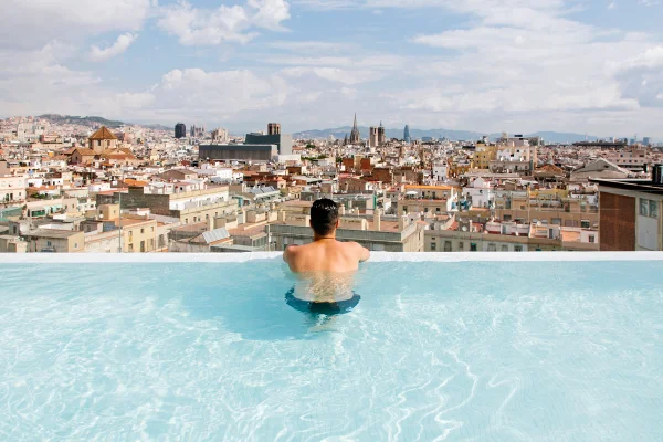 Um homem branco está de costas na beira da peiscina olhando para a cidade de Barcelona, na Espanha