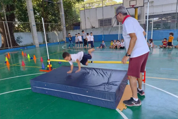 imagem colorida mostra criança saltando em cima de vara, em quadra de escola pública. um homem adulto acompanha a cena ao lado e outras crianças observam a cena ao fundo da quadra - metrópoles