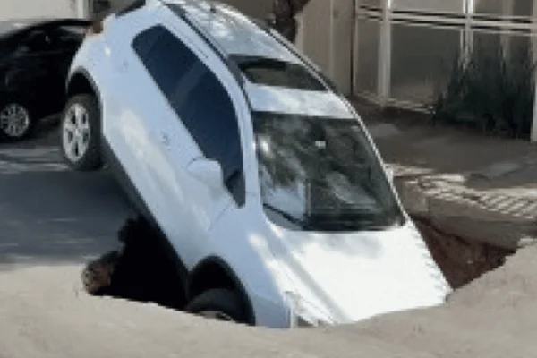 Imagem colorida de um carro branco engolido por um buraco após o asfalto ceder - Metrópoles