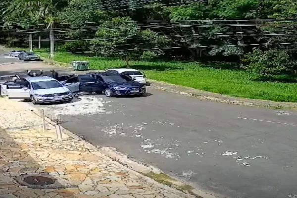 imagem colorida mostra assalto filmado por câmera de segurança. criminosos saem de um carro preto e rendem passageiros de carro de cor clara