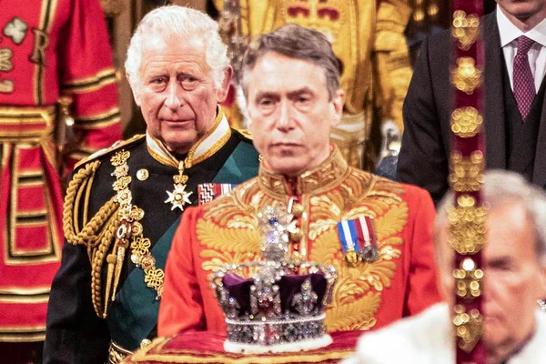Foto colorida. Príncipe Charles ao lado de uma coroa