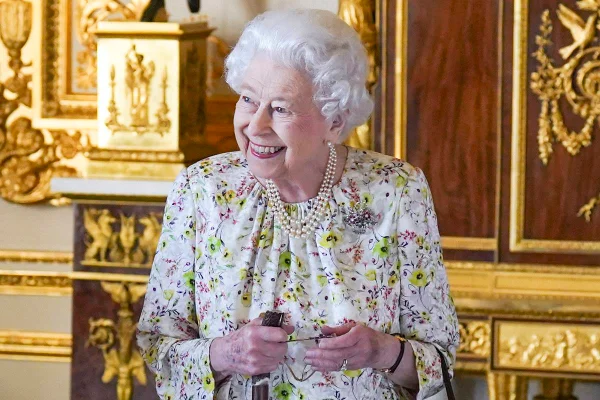 Foto colorida. Rainha Elizabeth com vestido florido, pérolas e uma bengala