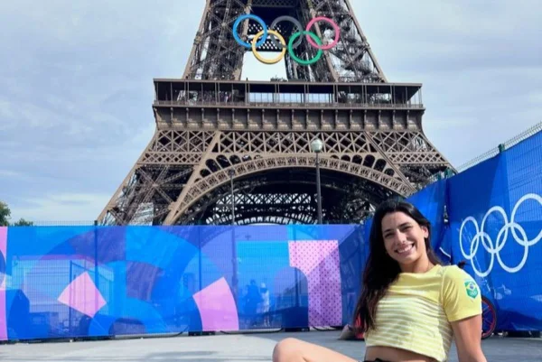 Imagem colorida mostra mulher branca com cabelos escuros longos e lisos. Ela veste camiseta amarela com listras brancas. Ao fundo está a Torre Eiffel com arcos olímpicos pendurados nela - Metrópoles