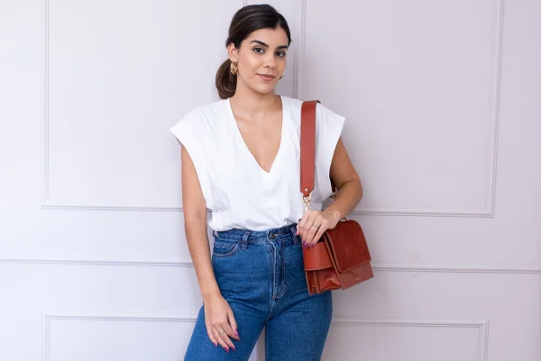 Marina Naves, mulher branca de cabelo curto preto amarrado. Ela usa uma camiseta branca, calça jeans azul e uma bolsa marrom de tiracolo