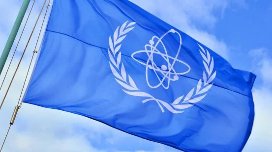 Bandeira da Agência Internacional de Energia Atômica, azul com representações de um átomo em branco, tremula no céu claro - Metrópoles