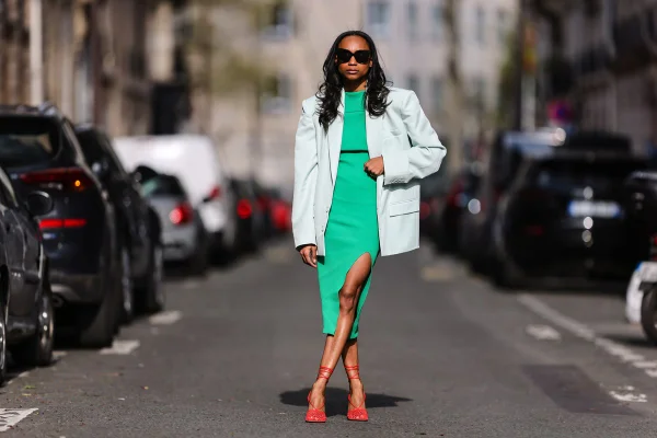 Look de street style: mulher negra usa vestido verde com fenda; blazer oversized cinza claro; óculos escuros; e sandália vermelha de salto