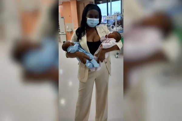 Imagem colorida de mulher em pé enquantoi segura dois filhos no colo - Metrópoles