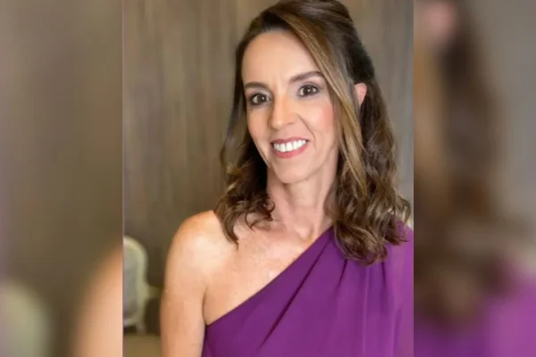 Imagem colorida mostra mullher branca com cabelos castanhos olhando para a câmera e com sorriso mostrando os dentes. Ela veste um vestido de festa roxo e tem os cabelos meio presos.
