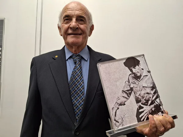 Imagem colorida mostra o capitão reformado da PM Augusto Carlos Cassaniga, um idoso calvo com cabelos brancos ao redor da cabeça, vestido com um terno preto e uma camisa azul recebendo homenagem pelo trabalho no combate ao incêndio no Edifício Joelma, em 1º de fevereiro de 1974 - Metrópoles