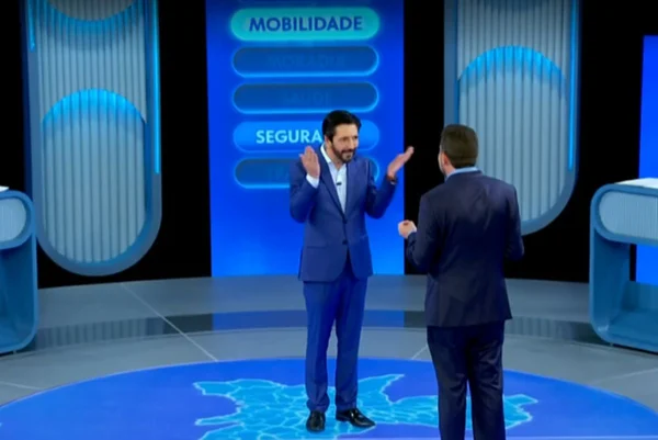 Imagens coloridas mostram um homem branco, de barba, com terno azul e camisa branca, olhando para outro homem em sua frente. Ele esá com os braços abertos.