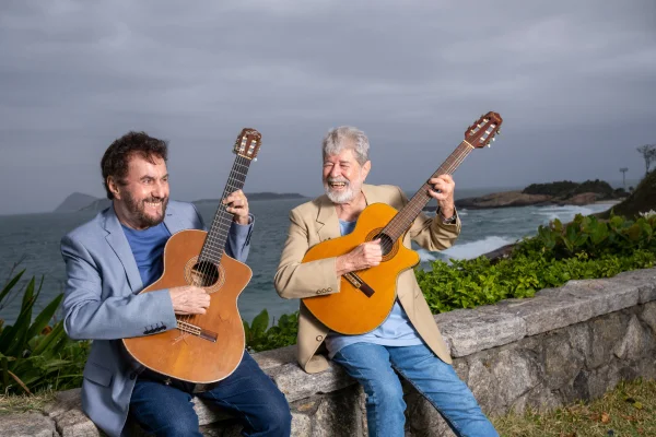 Fotografia colorida mostrando dois homens com violão em sacada com mar atrás-Metrópoles
